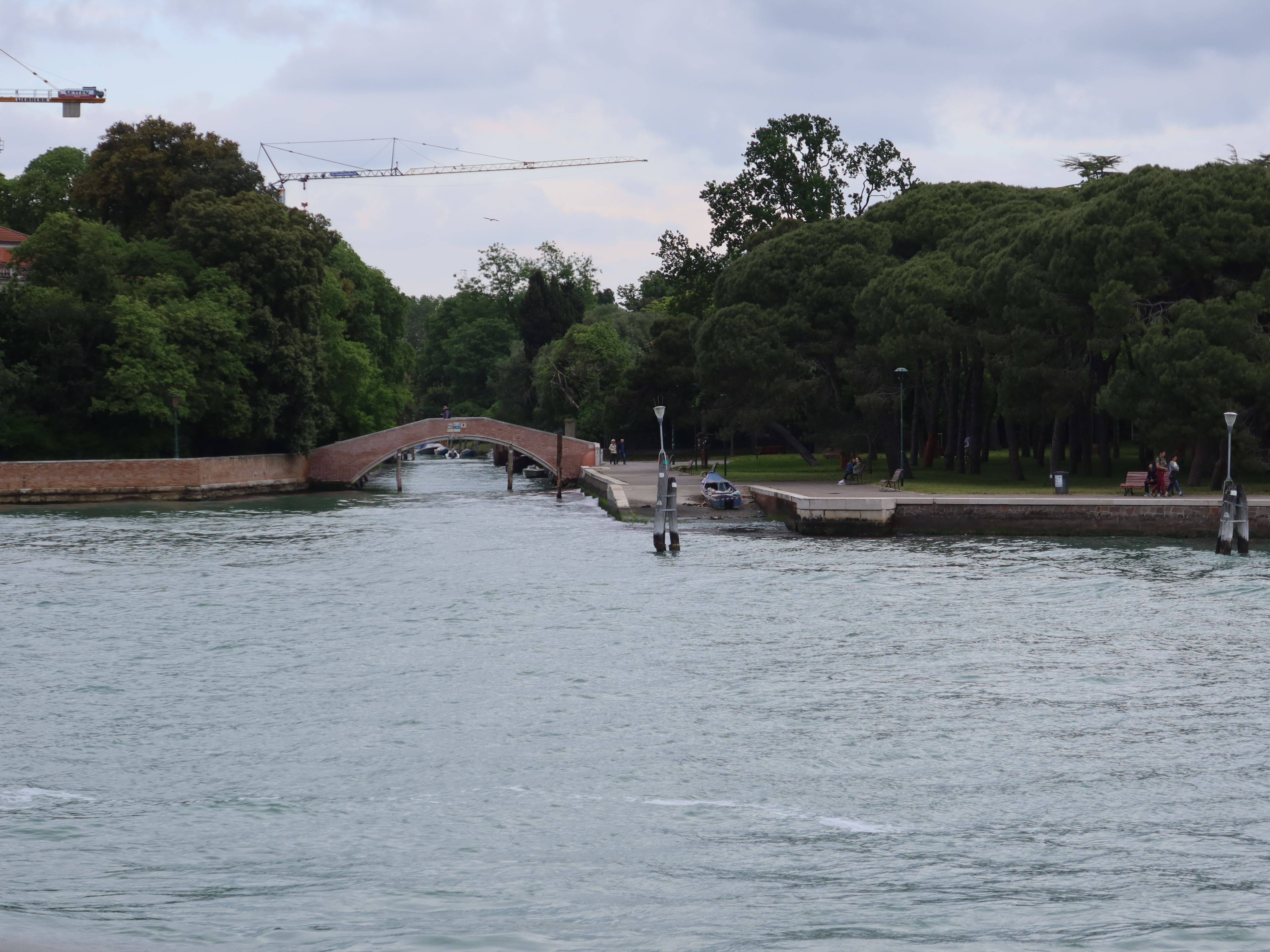 Brick Bridge Over Calm Canal Small arched brick bridge over canal with greenery and cranes under a cloudy sky.