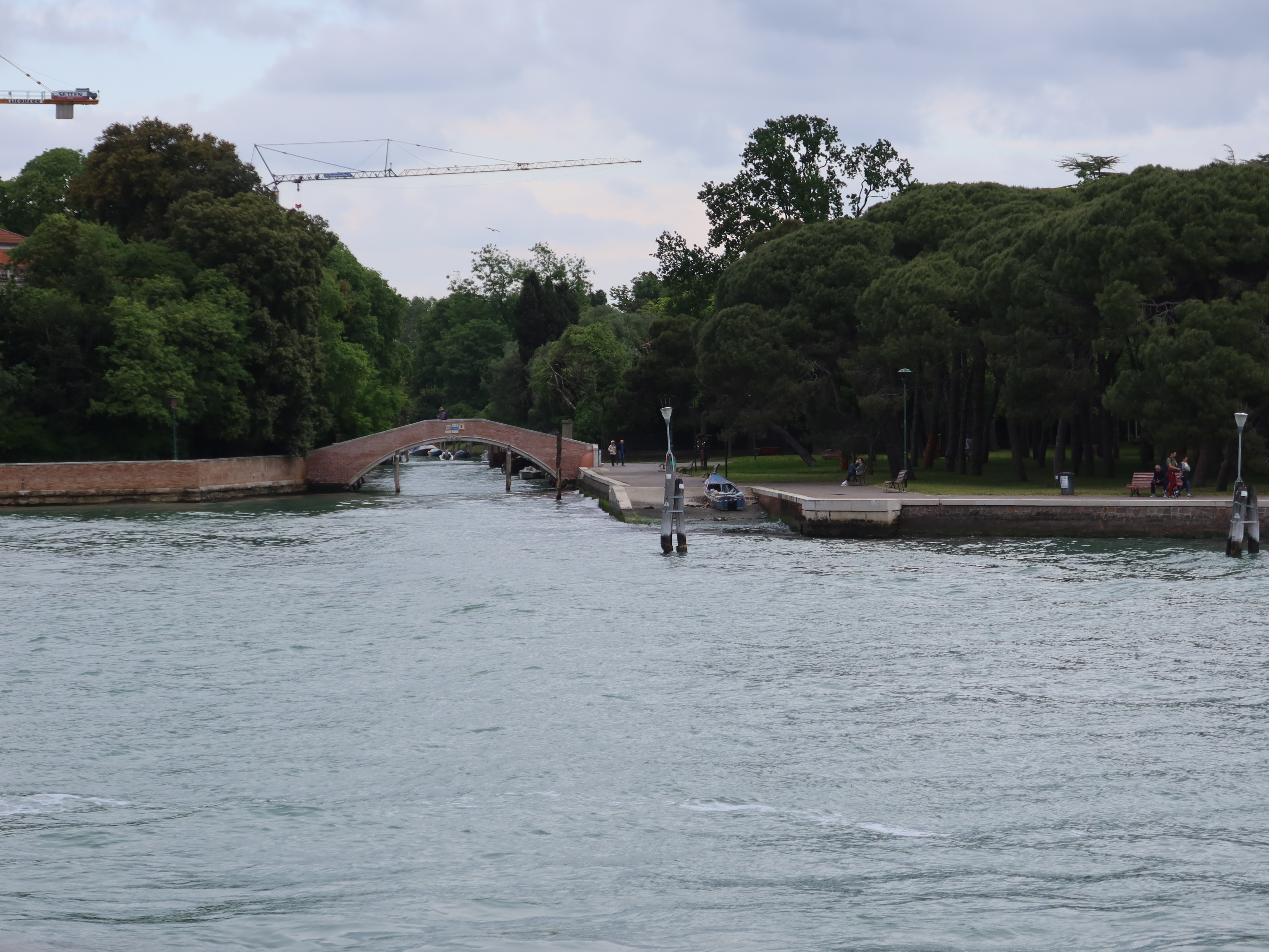 Waterway with green trees, arched brick bridge, path, cranes overhead, overcast sky.