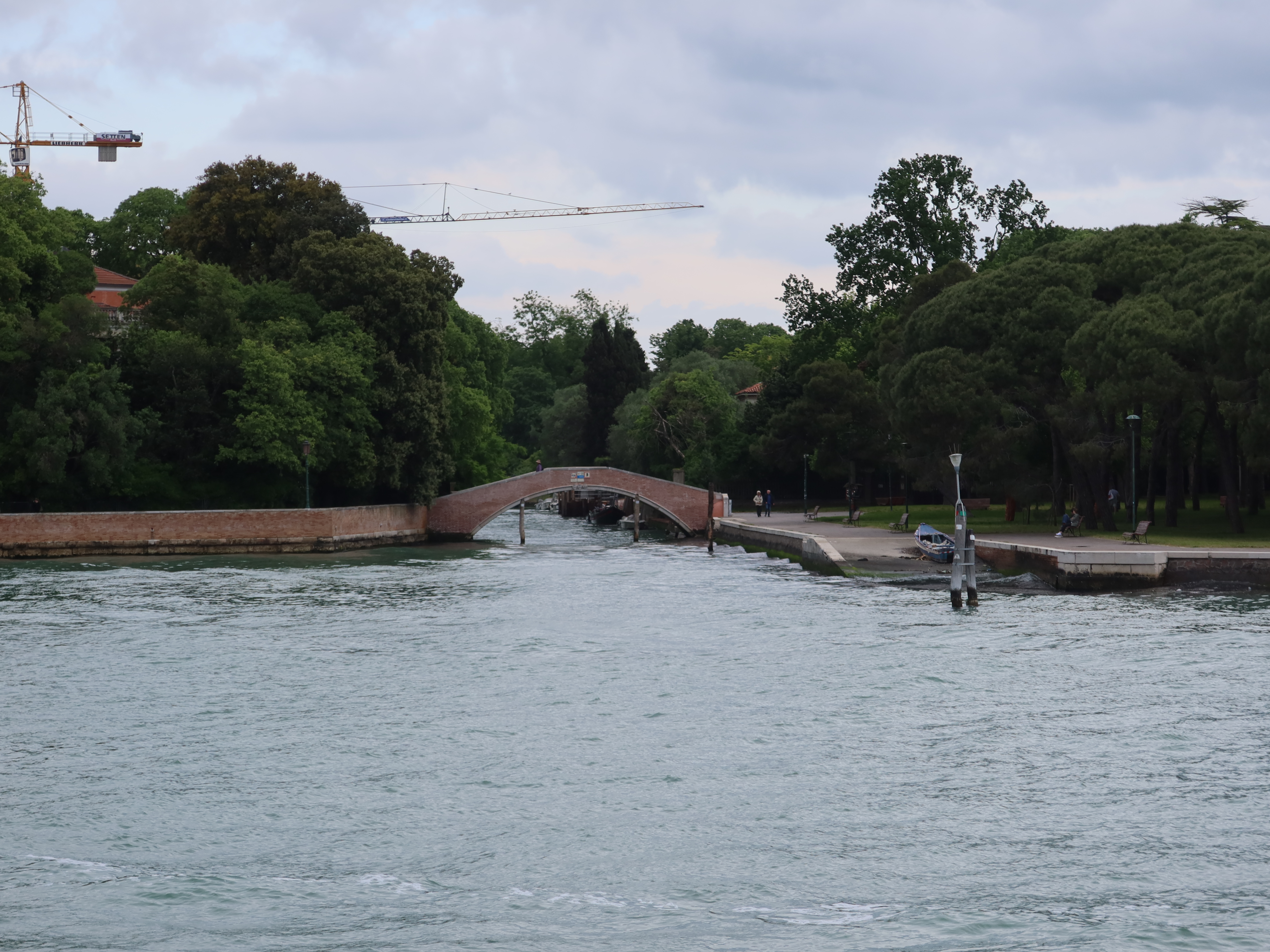 Brick Arch Bridge Over Canal in Tranquil Setting Brick arch bridge over canal with greenery, pathway, people, and crane in overcast sky.