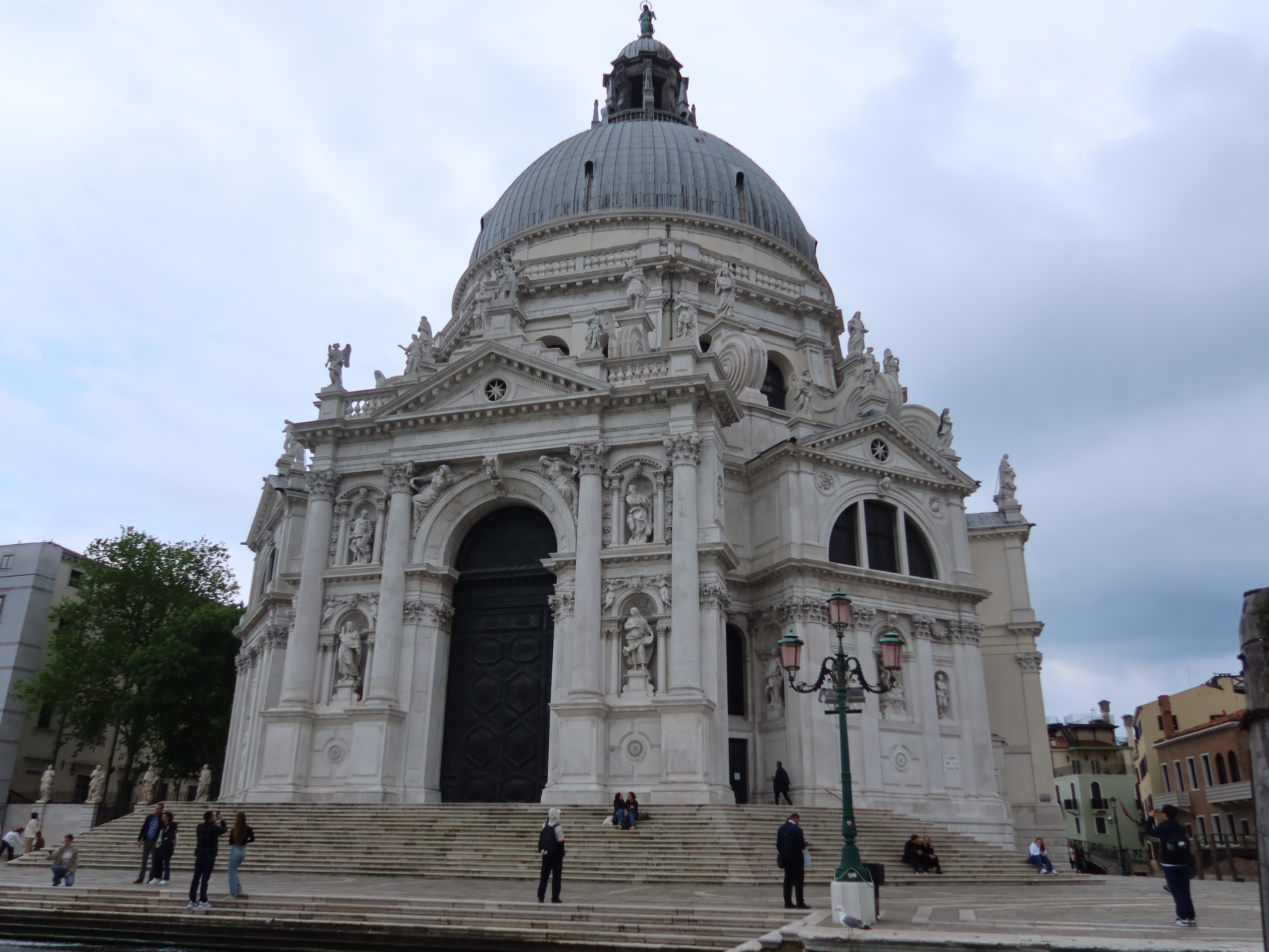 Ornate white church with dome, columns, statues, staircase; people on steps, cloudy sky.