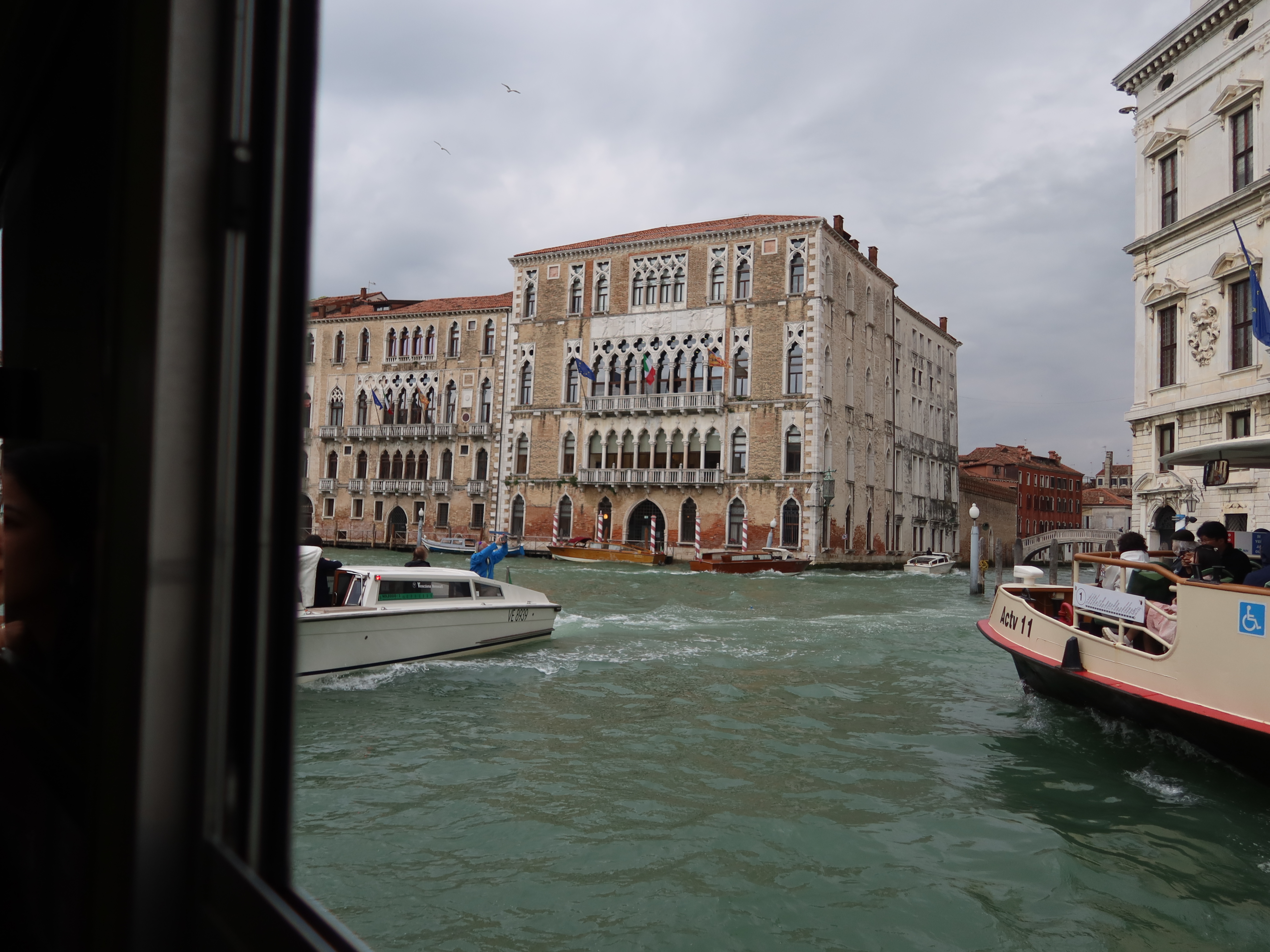 Historic building with flags by Venice canal, boats in water, overcast sky, seagulls above.