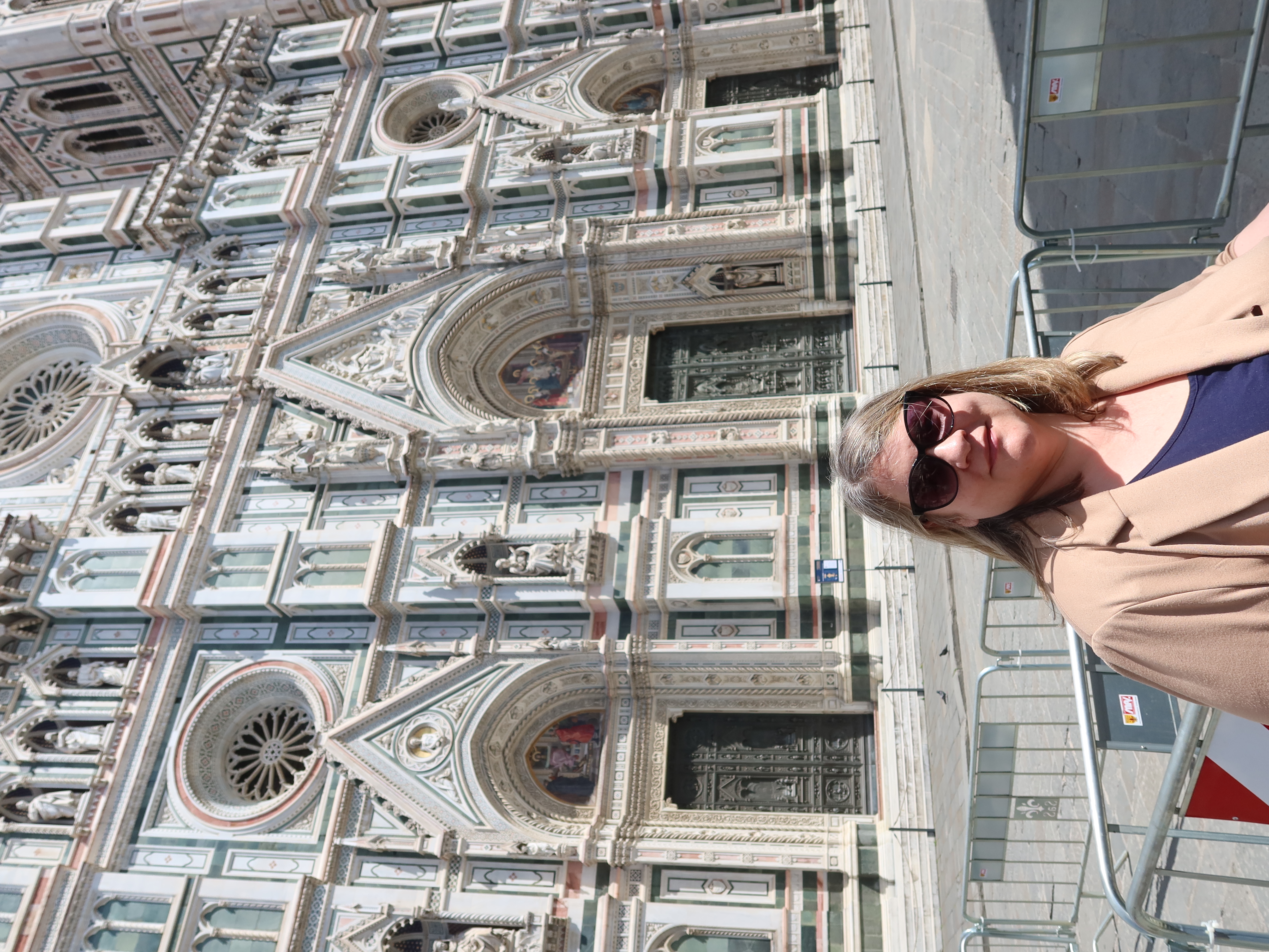 Woman in sunglasses and beige coat by Gothic cathedral with sculptures and large round windows.