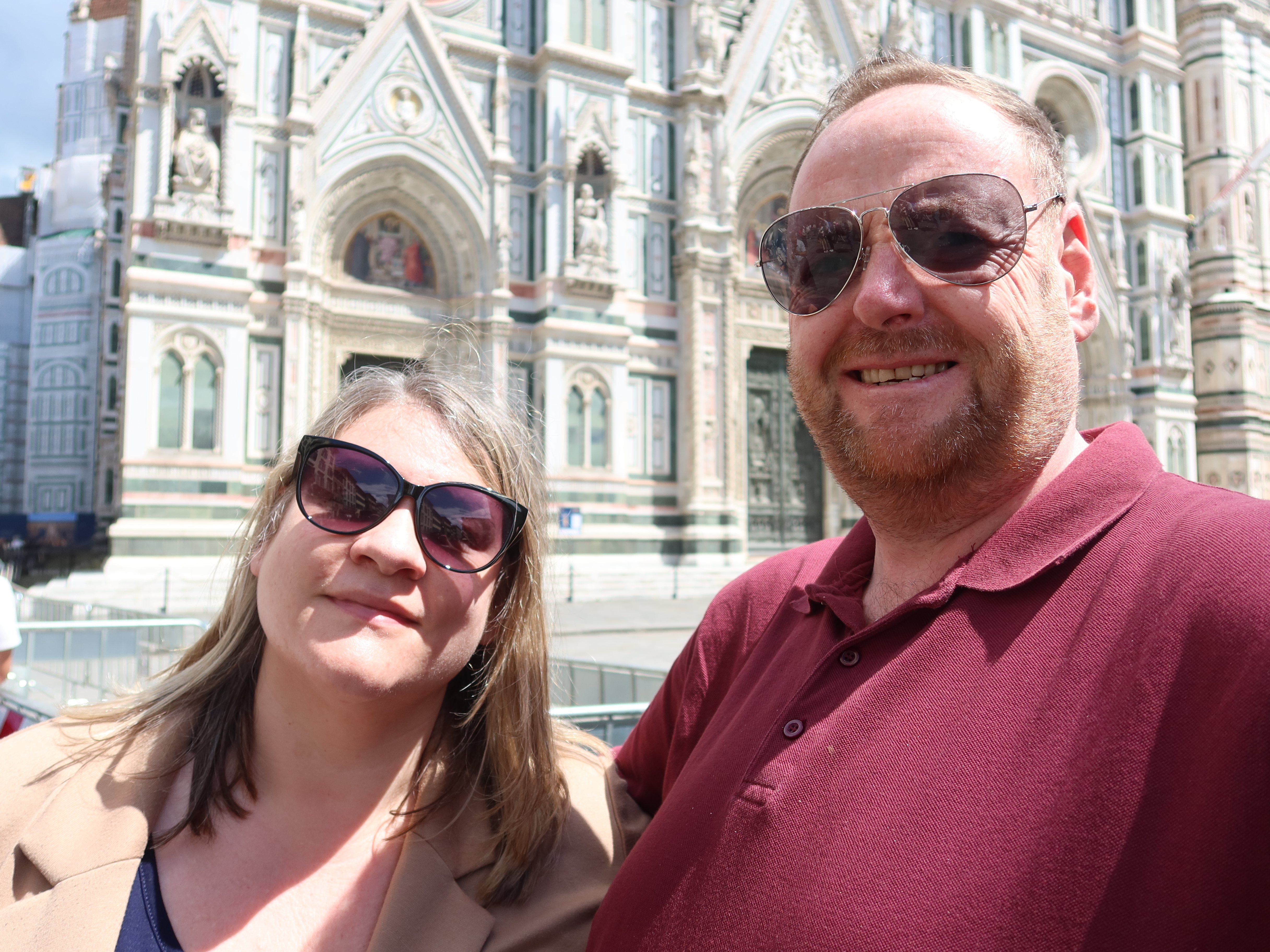 Man and woman in sunglasses smiling for selfie near ornate historical building with sculptures.