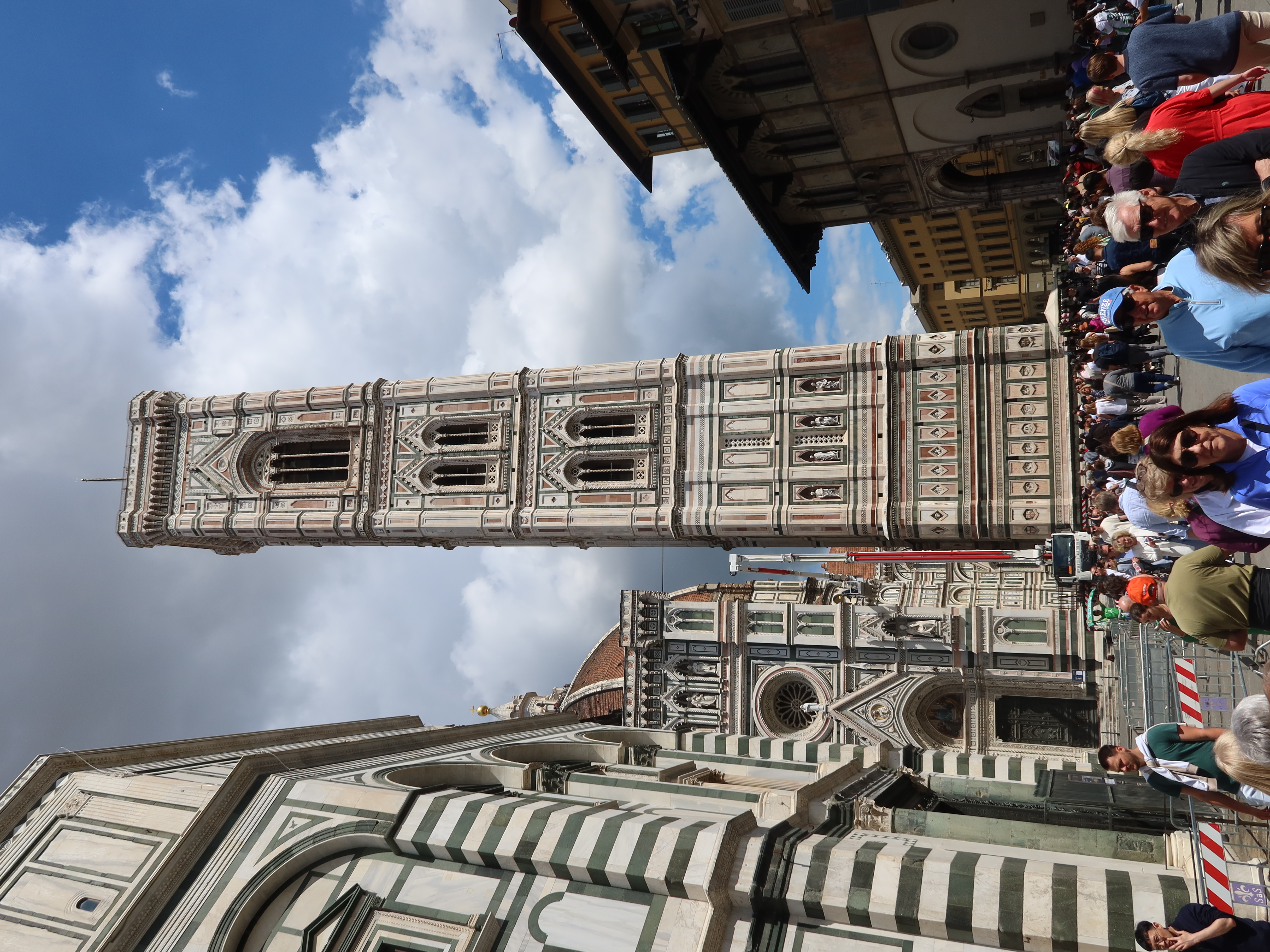 Ornate bell tower in busy urban scene, historic buildings, partly cloudy sky, crowd below.