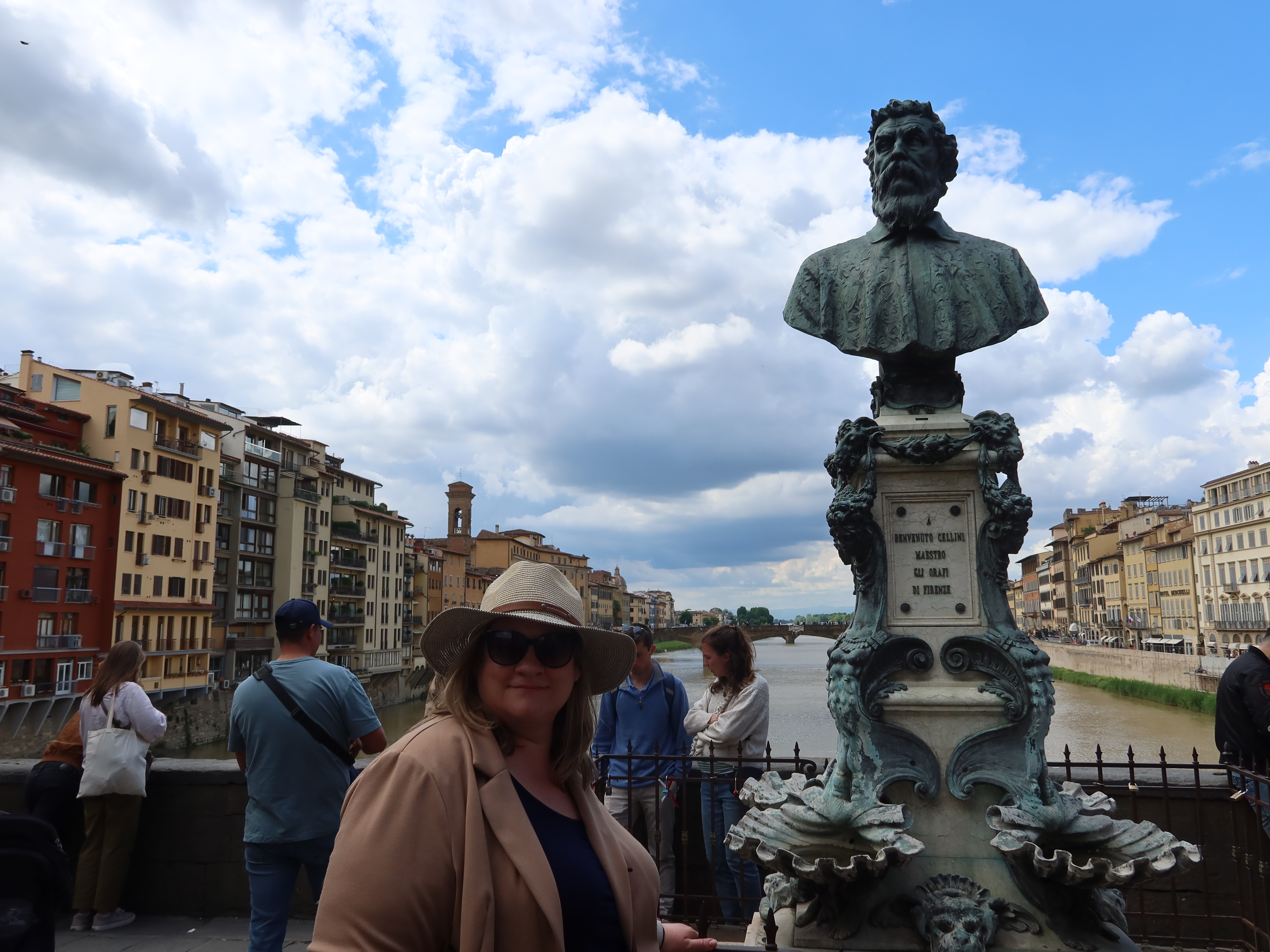 Woman near bronze bust on bridge, historic buildings and river in background, partly cloudy sky.