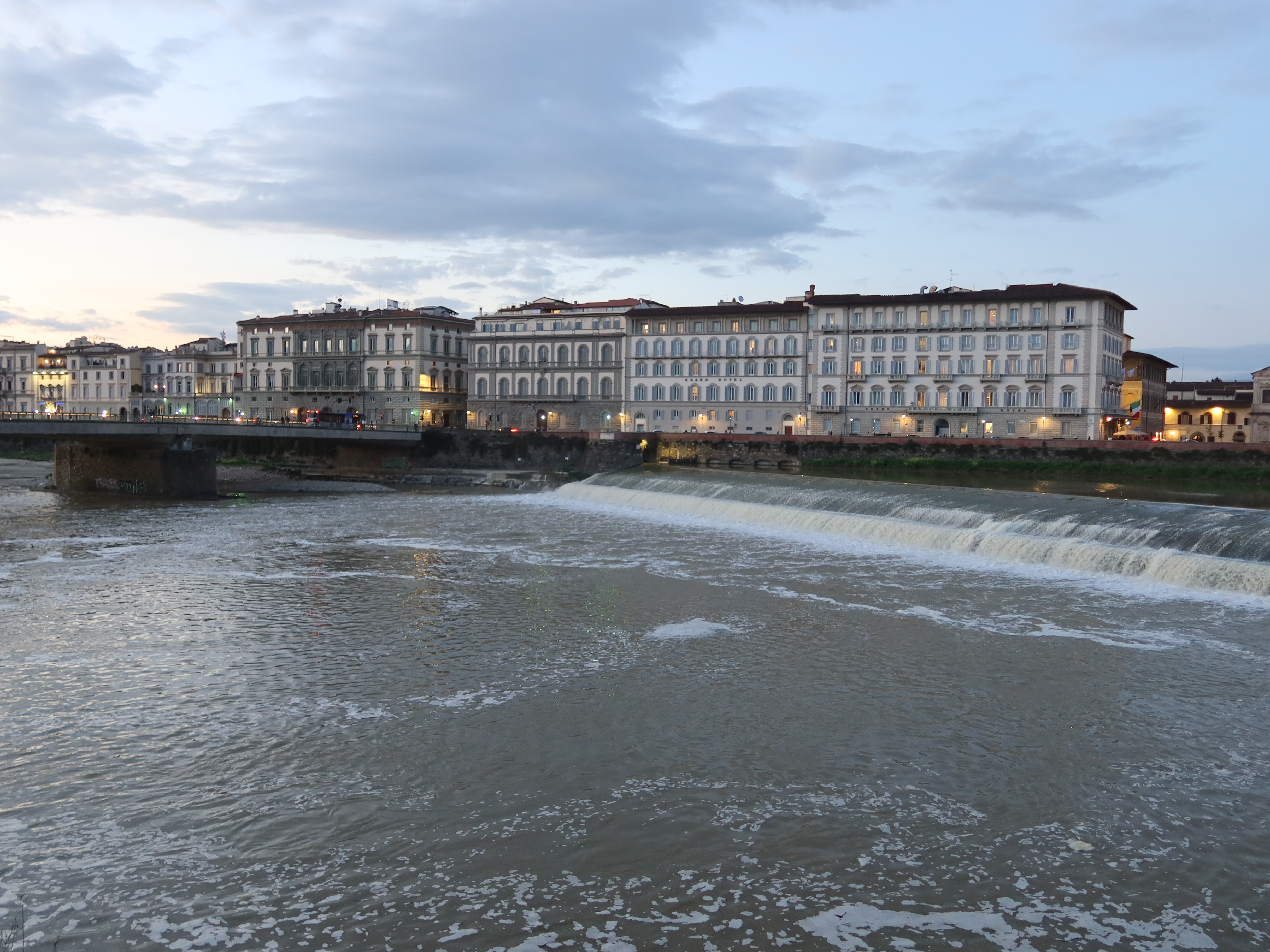 Wide river flows under bridge in urban area; buildings with lights and small waterfalls visible.