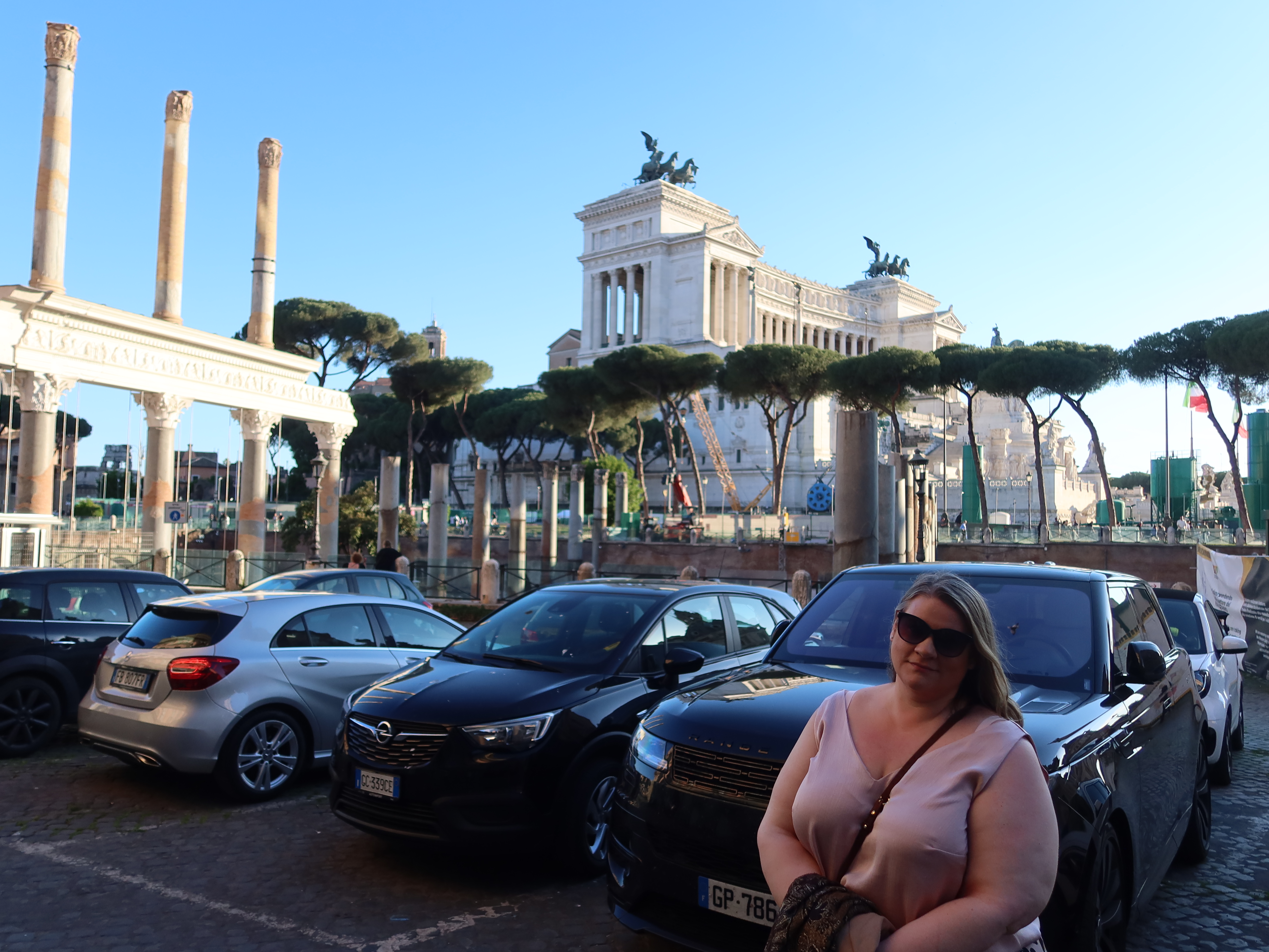 Person stands by parked car, historic building with statues in background, sunny day.