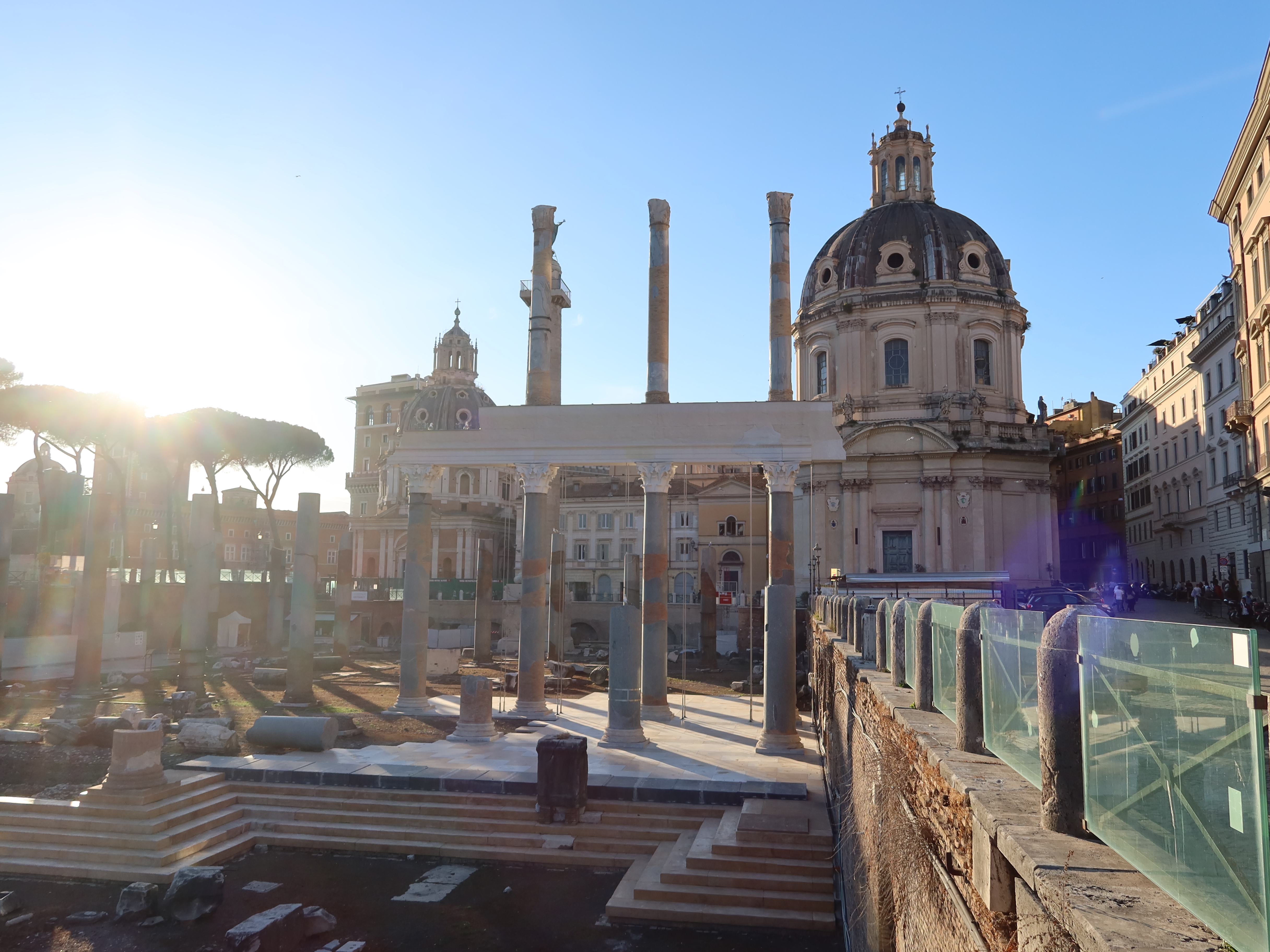 Ancient Roman forum ruins with columns and dome-shaped building at sunset in background.