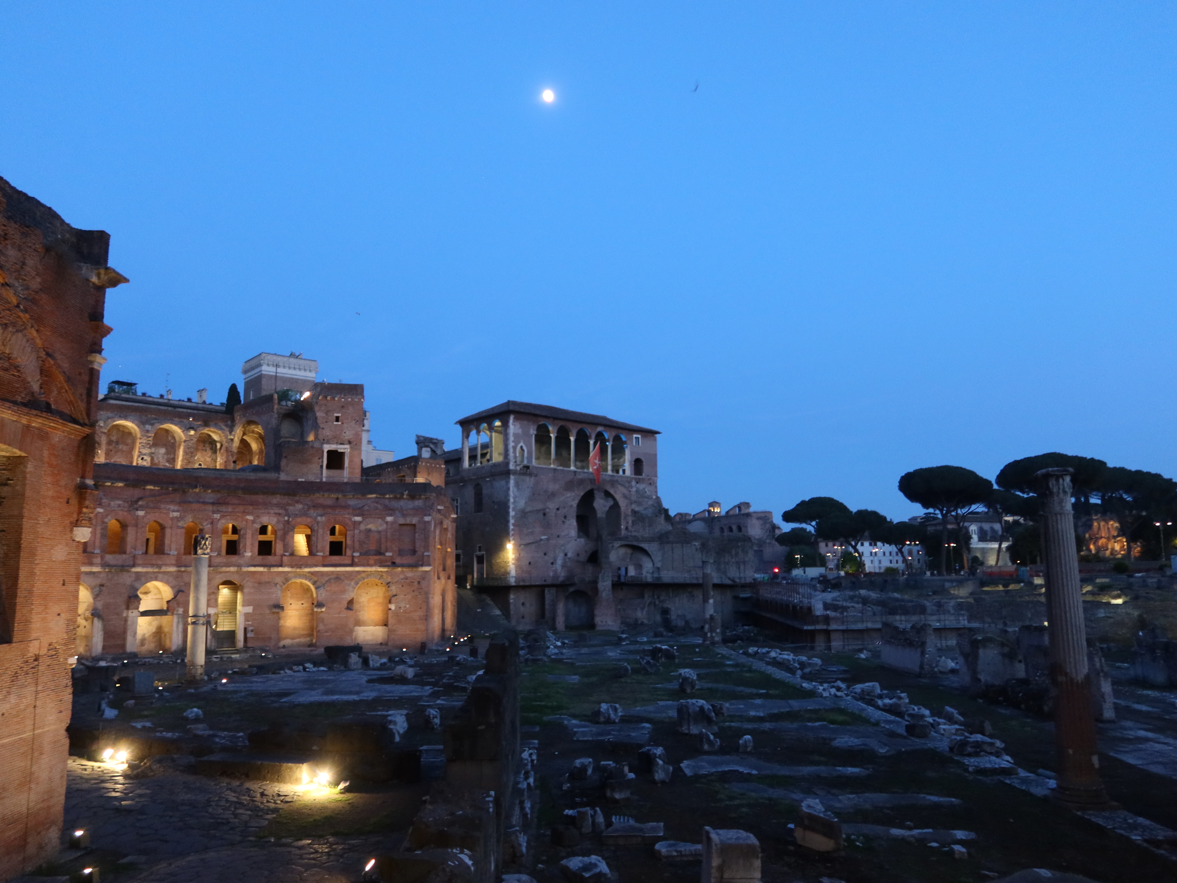 Ancient Roman ruin at dusk with arched windows, columns, ground lights, and moonlit sky.