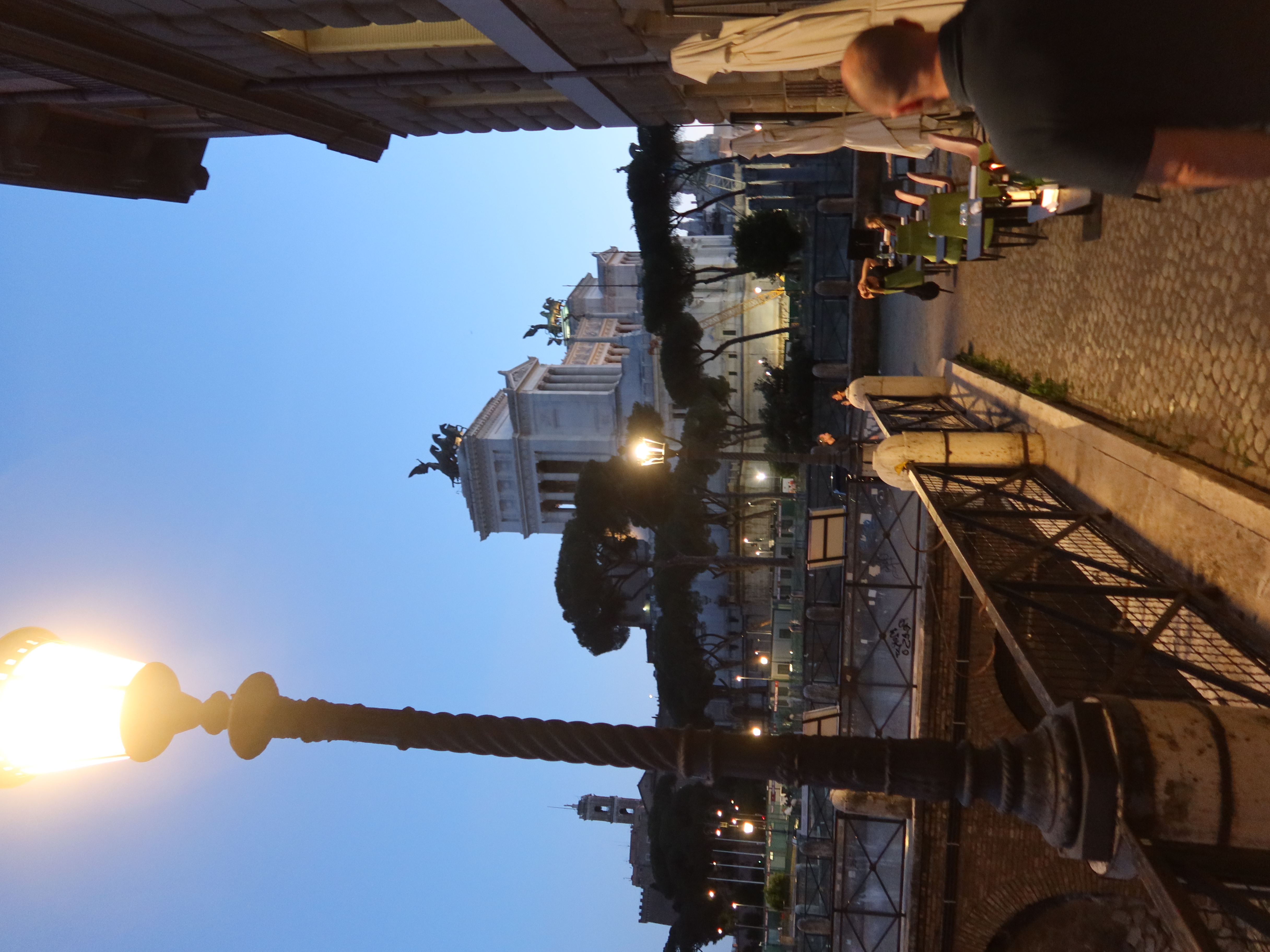 Street at dusk in European city; cobbled path, outdoor seating, lamppost, classical monument.