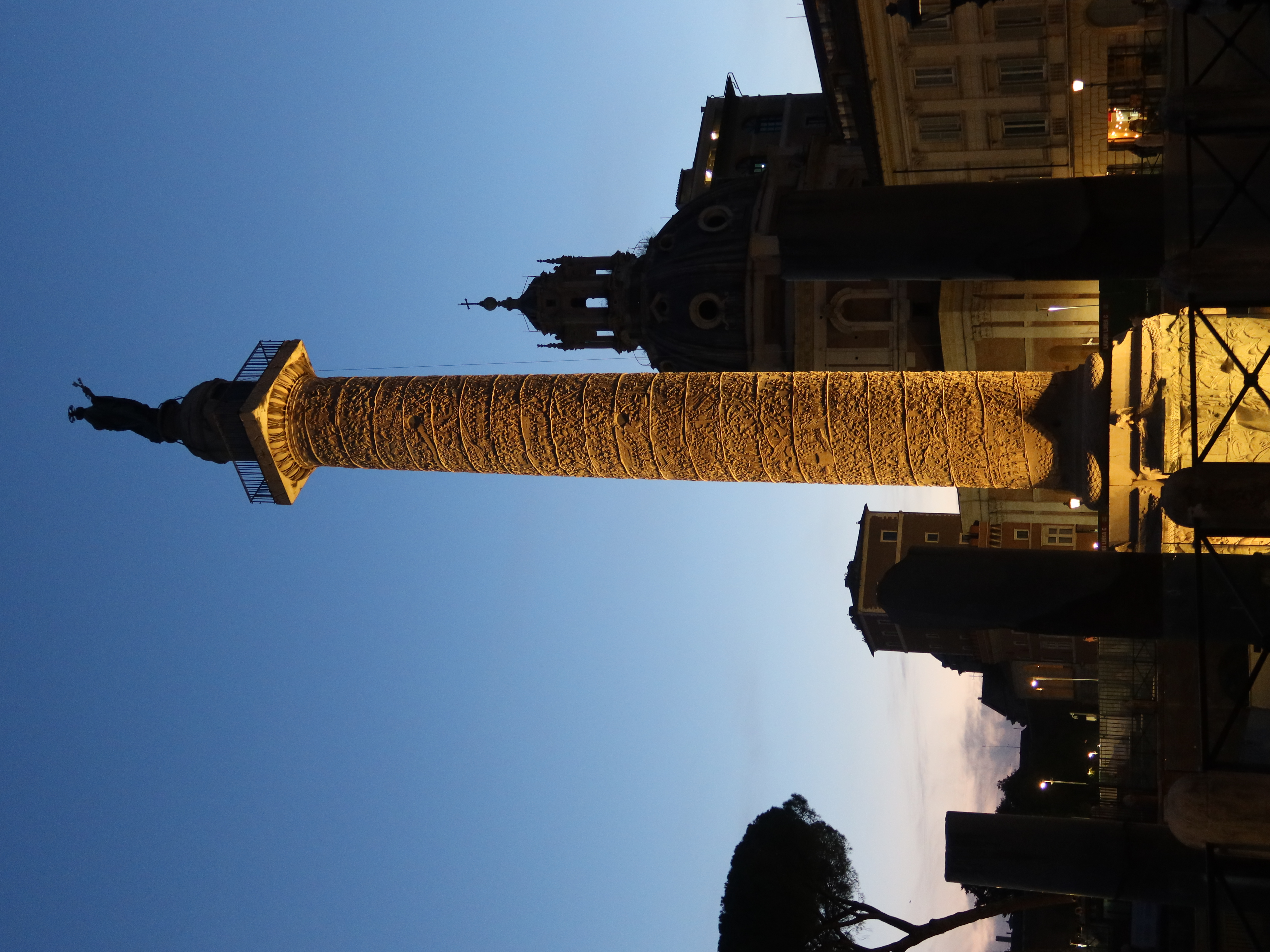 Tall carved column with statue, domed buildings, and tree silhouetted against twilight sky.