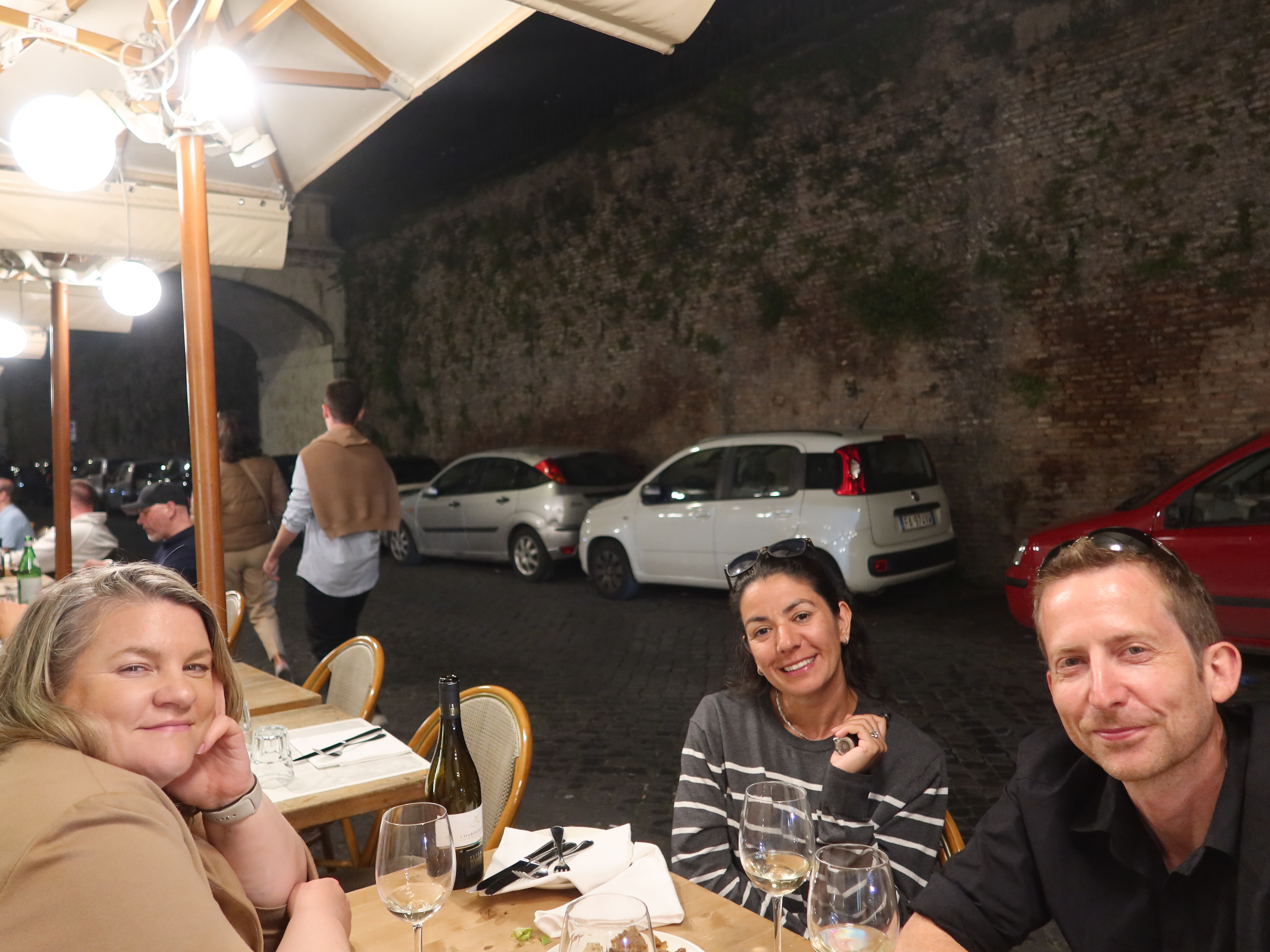 Three people dining outdoors at a restaurant table with wine, evening setting, cars in background.