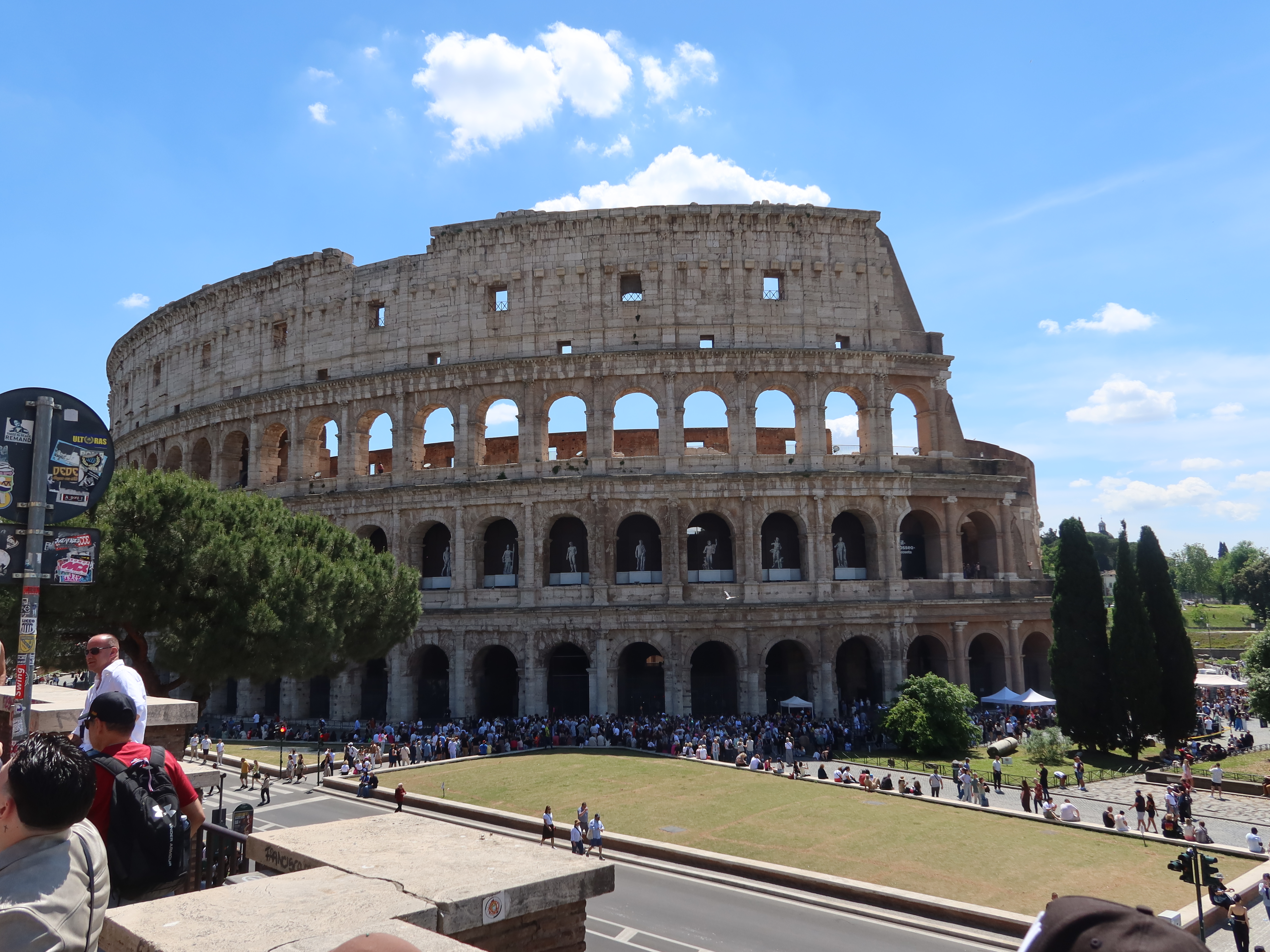 Colosseum in Rome with visitors, tree, signpost, blue sky, and partial stone arches visible.