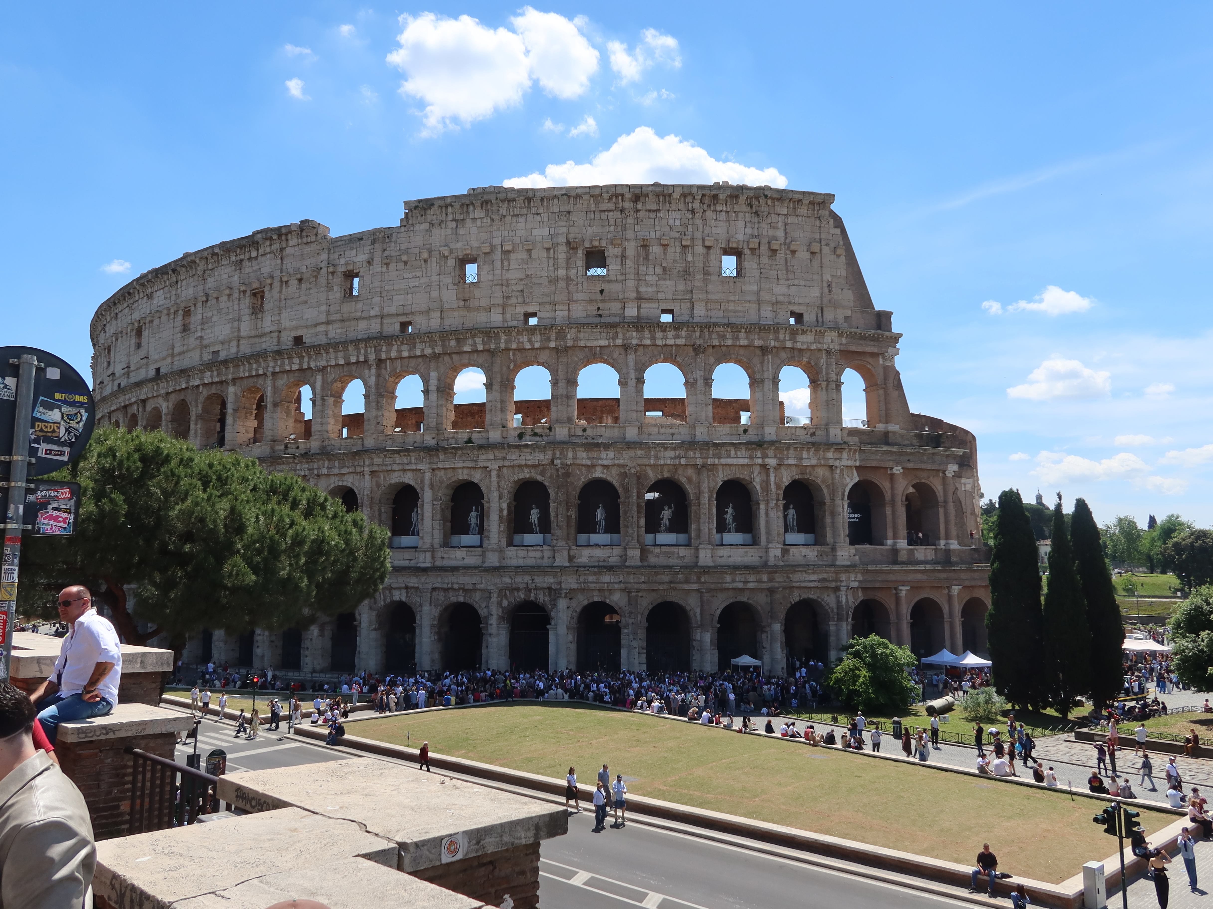 Colosseum Amidst Tourists on a Sunny Day Ancient Colosseum with tourists, arched levels, trees, clear sky, and nearby road in foreground.