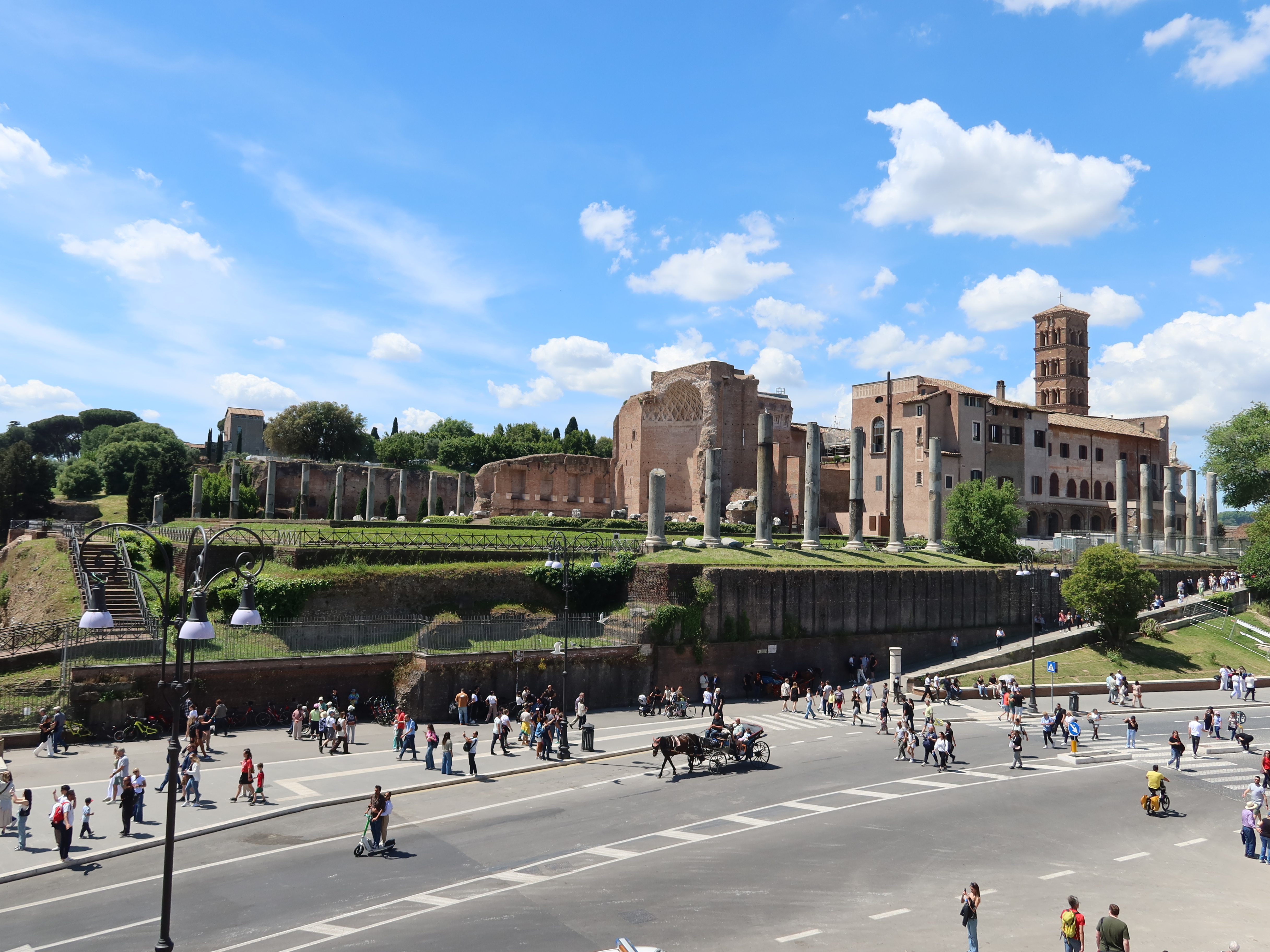 Ancient Roman ruins with columns, busy street, people walking, bicycles, horse carriage, clear sky.
