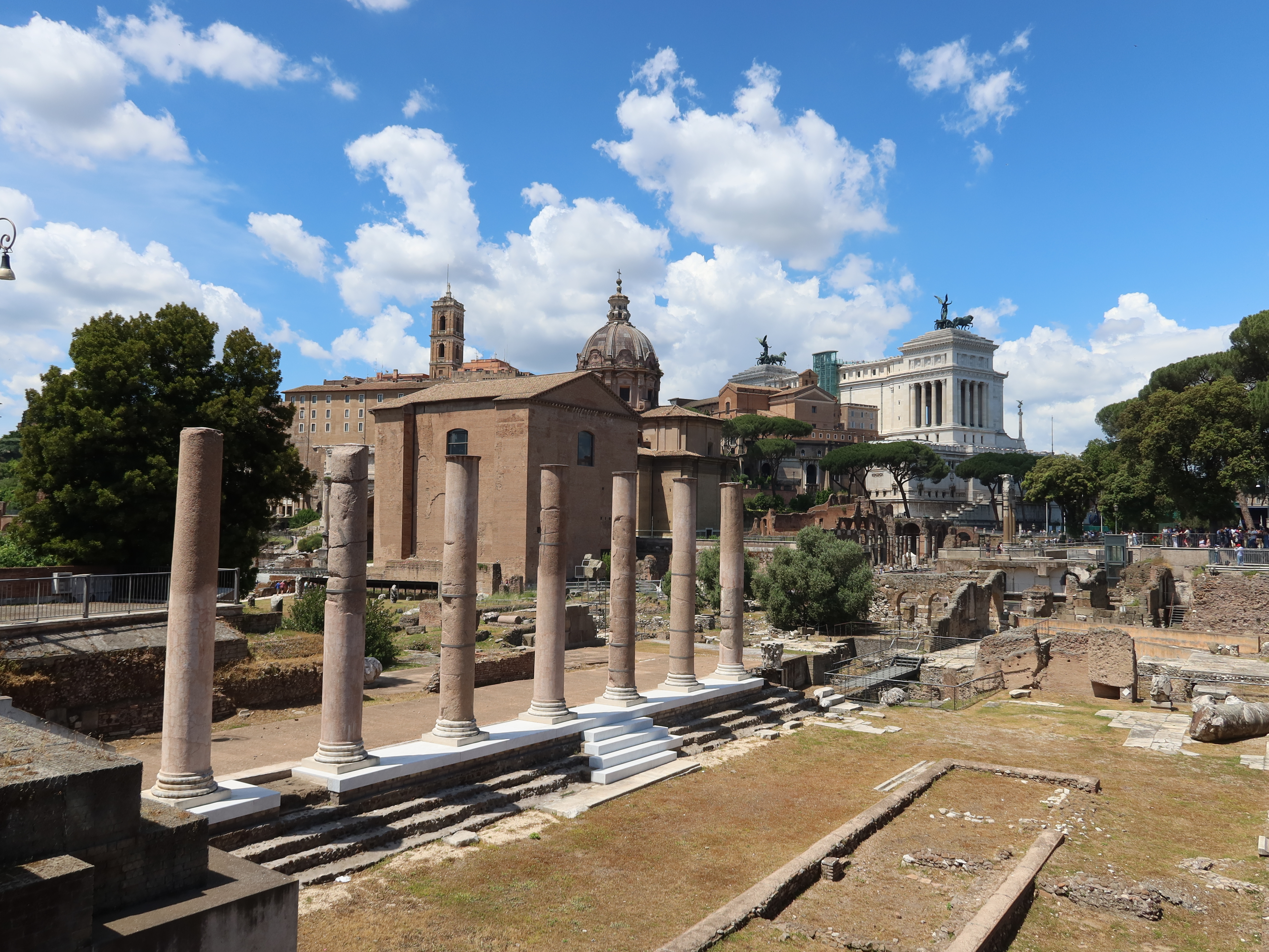 Ancient Ruins Under Blue Skies Ancient stone columns and ruins with classical buildings under a blue sky with clouds.